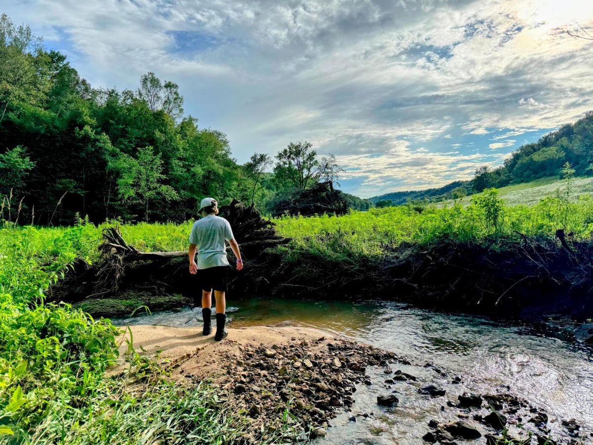 A child in rubber boots on a gravel bar in Biser Creek, with meadow and wooded ridges beyond