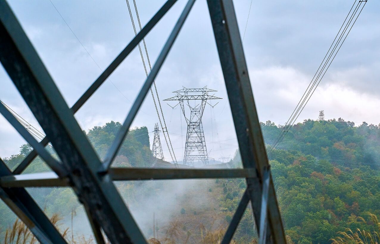 High-voltage lattice towers and lines crossing wooded hills in mist, seen through the frame of a tower